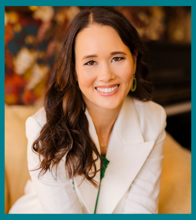 A professional headshot of a woman with long dark wavy hair wearing a white blazer. She has a bright, warm smile and appears to be in an indoor setting with a soft-focused background featuring warm tones.