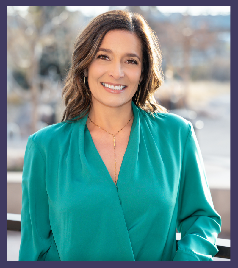 Alt text: A professional headshot of a woman with shoulder-length brown hair wearing a vibrant turquoise wrap blouse and a delicate gold necklace. She is smiling warmly at the camera with a bright, genuine expression. The photo appears to be taken outdoors with a soft, blurred background.